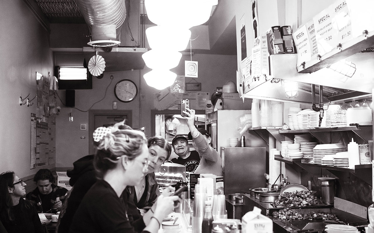 A black and white image of the backside of a crowded diner with a bar to the right and sets of small tables lining the wall to the left. The counter of the bar is filled with breakfast foods, cups, and dishes. In the background, a man and a woman are taking a selfie on their phone. 
