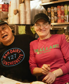 A feature image of a man and a woman standing behind a diner counter and smiling wide for the camera. The man has his arm around the woman and is wearing a black shirt with red and blue lettering and a black baseball cap. The woman is wearing a pink shirt with green lettering and the sleeves pushed up slightly. The woman is also wearing a gray baseball cap.