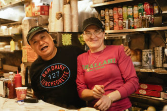 A feature image of a man and a woman standing behind a diner counter and smiling wide for the camera. The man has his arm around the woman and is wearing a black shirt with red and blue lettering and a black baseball cap. The woman is wearing a pink shirt with green lettering and the sleeves pushed up slightly. The woman is also wearing a gray baseball cap.