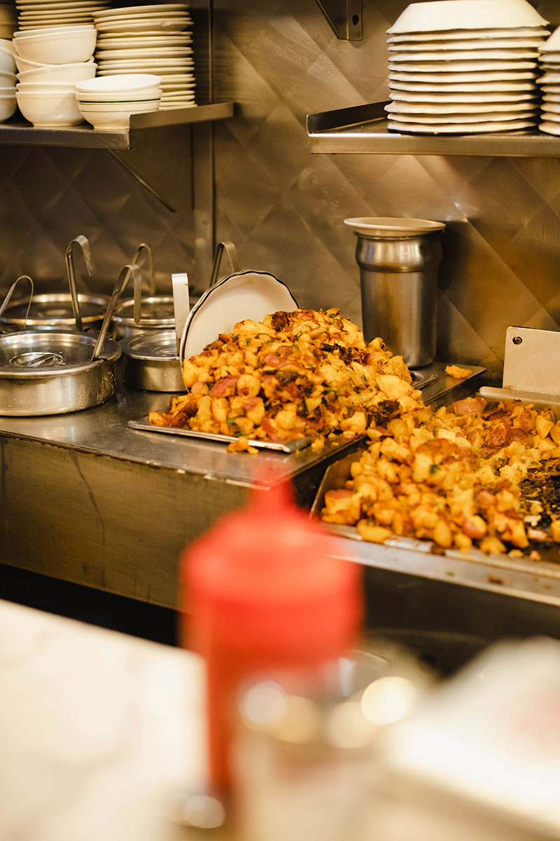 A photo of a griddle behind a diner counter filled with heaps of potatoes. Stacks of white bowls and plates are on the shelf above the griddle. A red plastic ketchup bottle is visible in the foreground of the photo.  