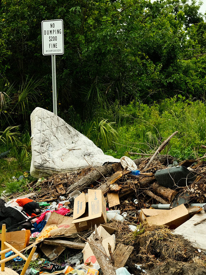 A photo of a large pile of random items and brush dumped on the side of the road. A large dirty white mattress is haphazardly resting against a black and white no dumping sign. Bright green trees and vegetation surround the area.