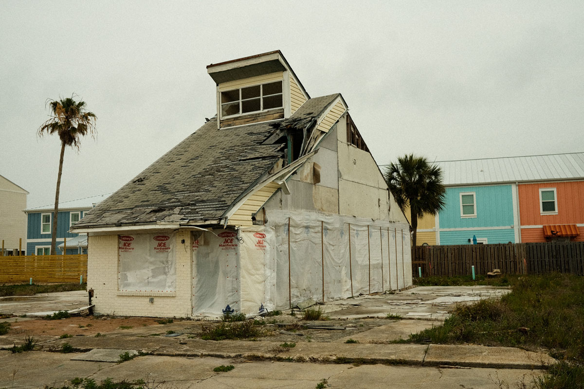 A photo taken on a gloomy, cloud-covered day of an old boarded up off-white house with a splintered black shingled roof. The side walks around the home are covered in cracks and overgrown weeds. A line of brightly colored, newly built, orange, yellow and blue condos sit contrastingly behind the old brown wooden fence in the background.A single palm tree is to the left of the old house.  