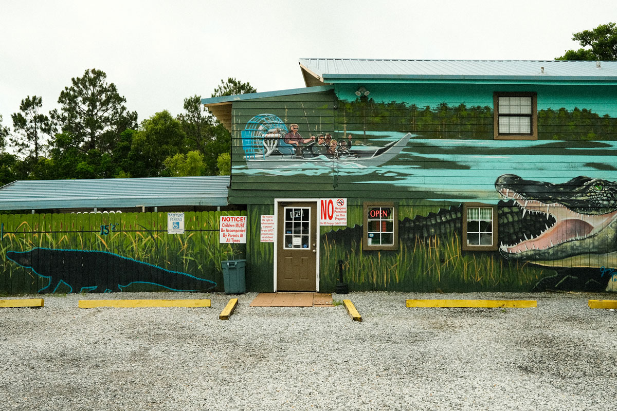 Photo of a wooden building and a gravel parking lot. The building is painted to look like a scene in the everglades with a man piloting a a fan boat in light turquoise waters covered with trees and vegetation towards the top of the buildings wall and a large swatch of bright green grass hiding a large, dark green alligator at the base of the building's wall. 