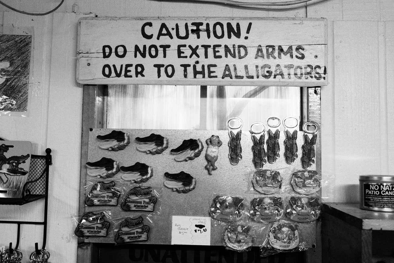 Black and white photo of a large, hand-painted wooden sign inside a shop and a metal board covered in alligator-themed souvenir magnets.