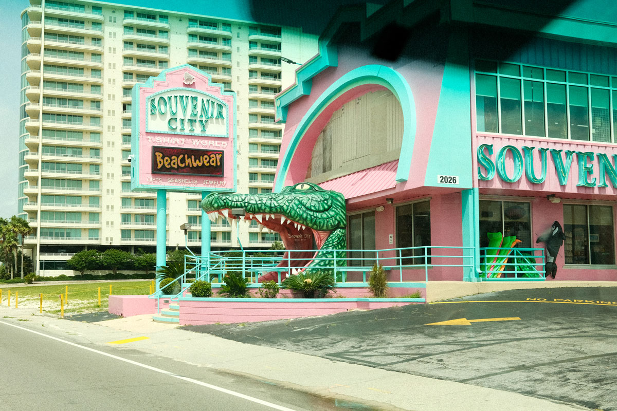 Photo taken from a car dashboard of the outside of a brightly bubblegum pink- and turquoise-colored building. The doorway to the building is through a large green alligator's mouth.Behind the building is is a multi-storied off-white apartment building.