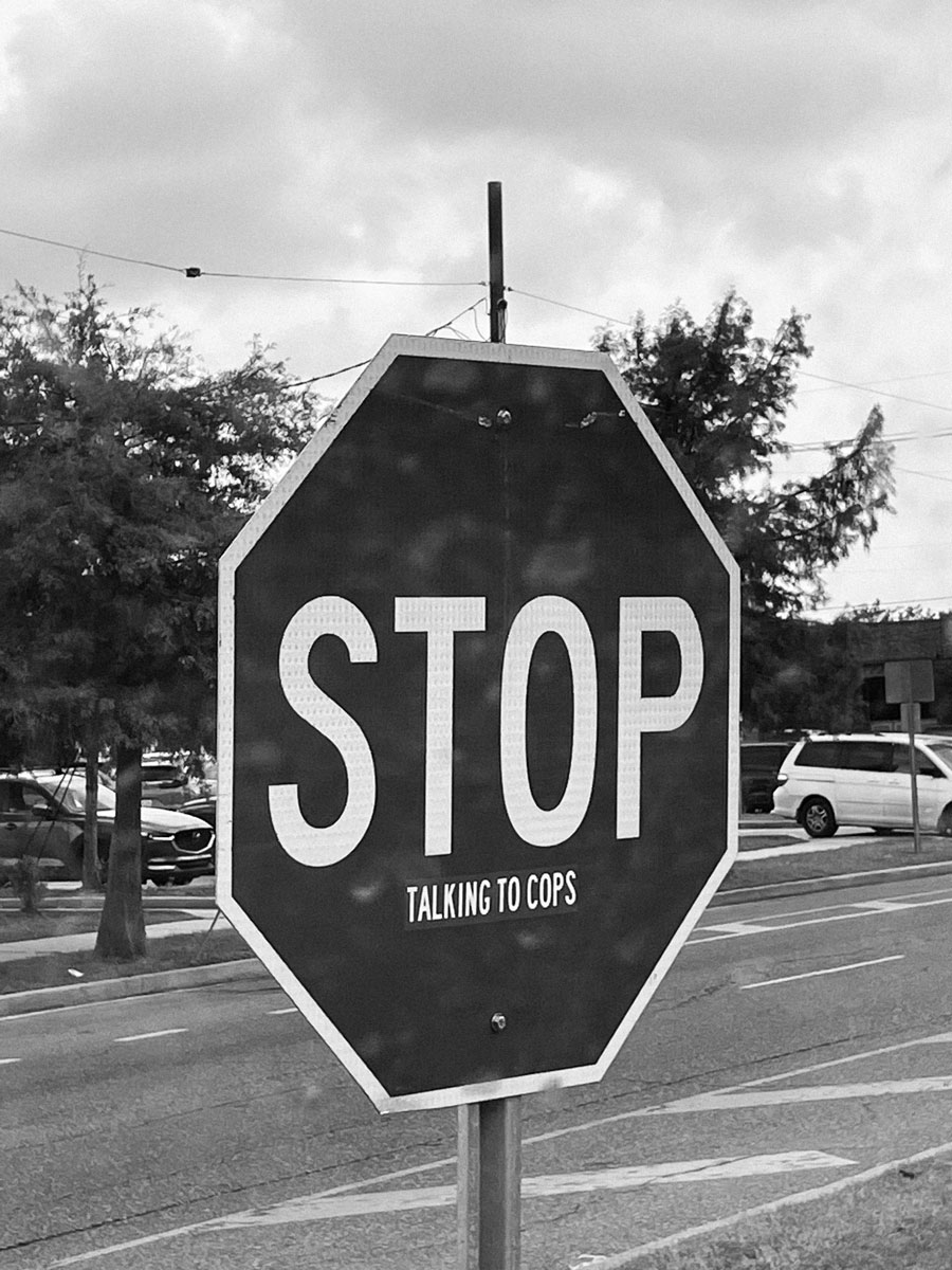 Close-up black and white photo of a stop sign with white lettering that reads "STOP" and smaller lettering beneath that reads "TALKING TO COPS".