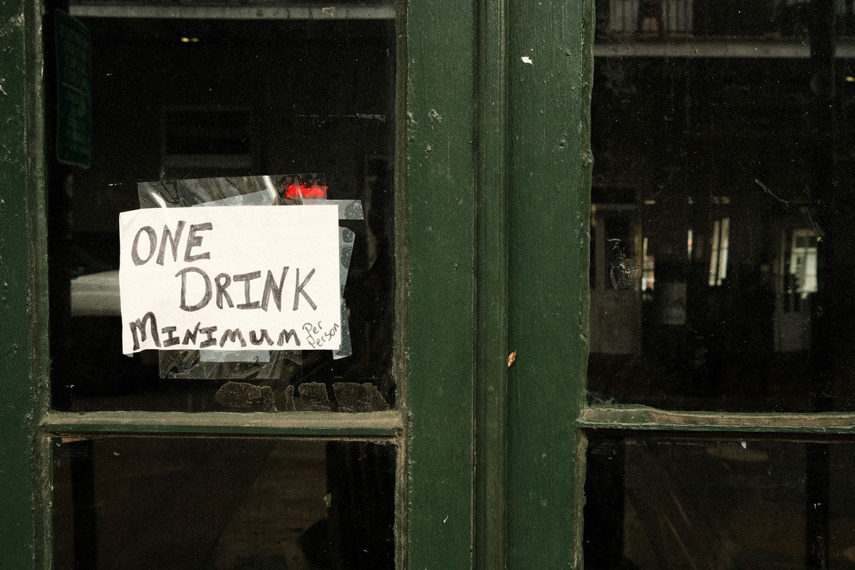 Photo of the dark green wooden shop doors with tinted windows and a small pieces of paper taped the left door with the words "ONE DRINK MINIMUM PER PERSON" handwritten in black marker.