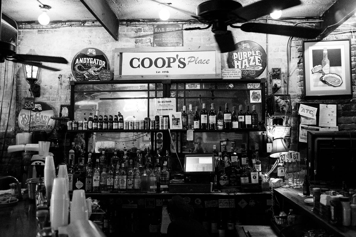 Black and white photo of the inside of a dimly lit, small dive bar with a mirror behind the bar and two shelves crammed with bottles of alcohol. The walls are brick and logos of various alcohol brands hang from them. Two large working fans hang from the ceiling. 