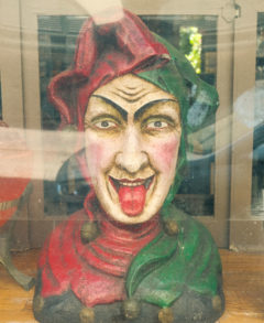 Photo of a fortune teller doll bust in a green glass case. The doll is dressed in a red and green jester's costume and is sticking its tongue out.