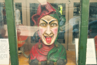 Photo of a fortune teller doll bust in a green glass case. The doll is dressed in a red and green jester's costume and is sticking its tongue out.