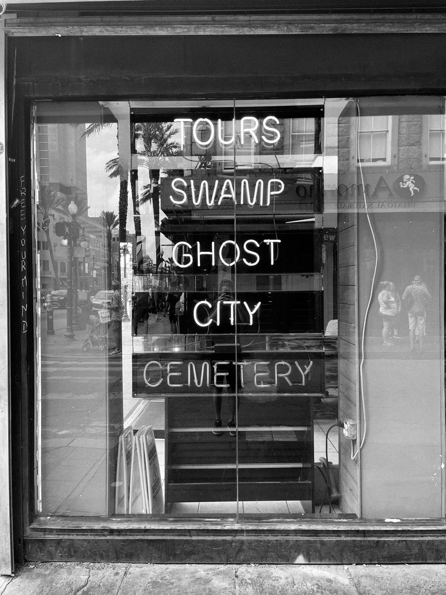 Black and white photo of a sidewalk shop window with 5 stacked LED signs advertising different types of tours hanging from the inside of the shop. 