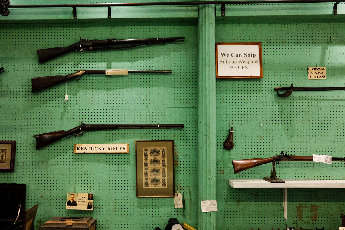 Photo of a jade green wall in the inside of an antique shop. The wall is covered in displays antique rifles for sale. 