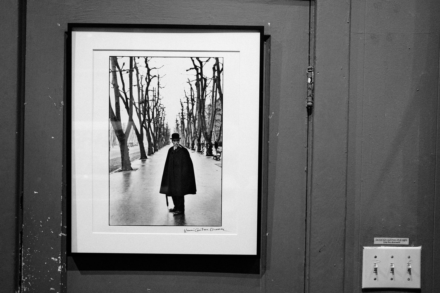 Black and white photo of a black framed photograph hanging from a painted wooden door. The vertical photograph is mounted on white paper and is of a man standing in the middle of an eerie and wet tree-lined street. The man is staring at the camera while a cigarette dangles from his mouth. He is wearing a suit under a black duster coat and a bowler hat and holding a closed umbrella. A small black signature is scrawled in the bottom right corner of the mounted paper.