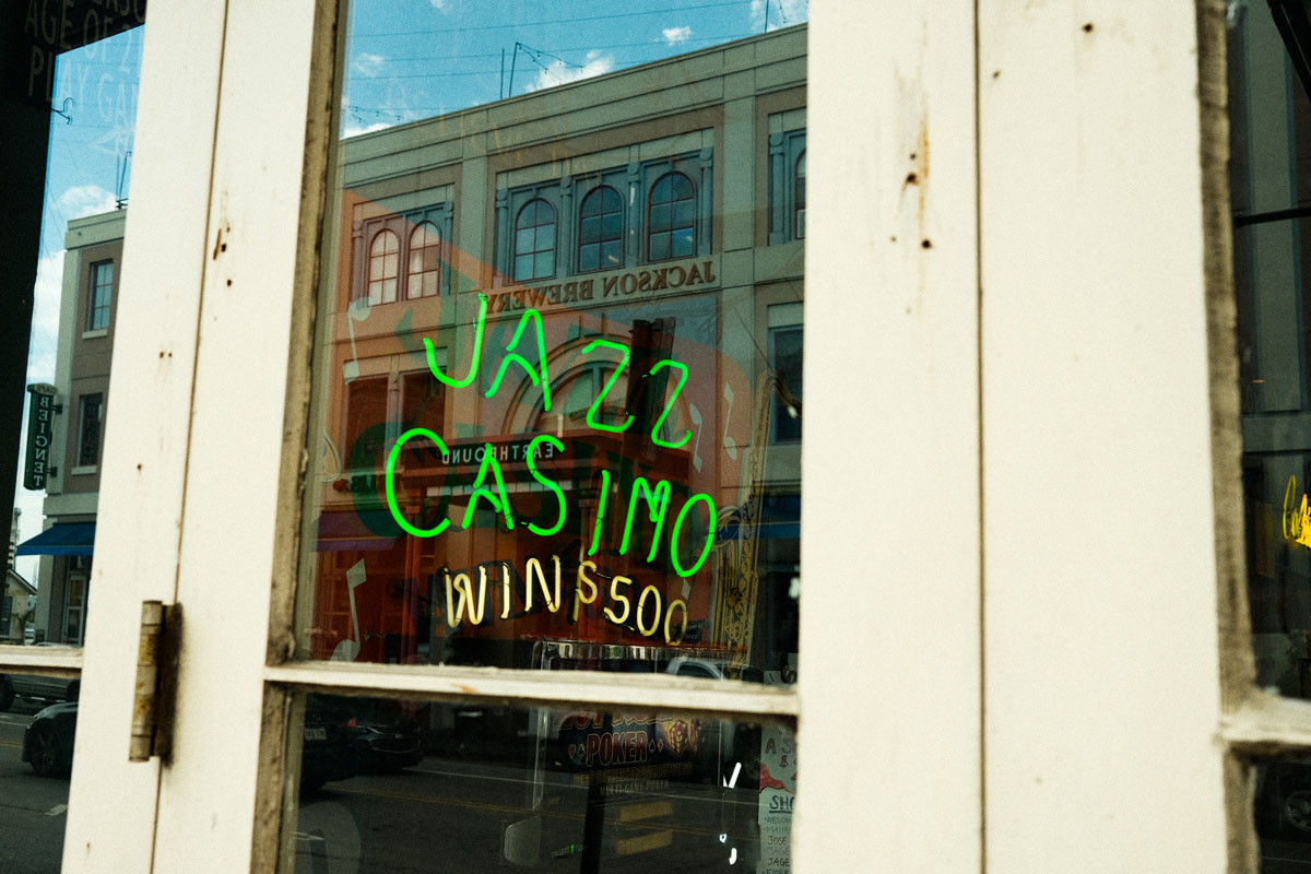 Photo of the outside of a shop with white wooden-lined glass doors. A bright green LED sign that reads the words "JAZZ CASINO" is hanging from the inside of the glass door. The reflections of large building across the street are visible in the window glass.