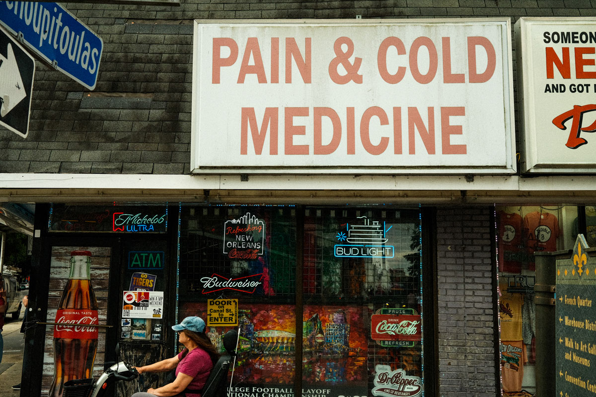 A small gray, brick sidewalk shop with a large, dirty white sign with faded red lettering. A colorful array of LED alcohol and drink signs cover the shop window from the inside and the door is wrapper in an image of a glass coca cola bottle.