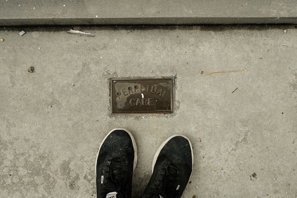 Photo taken by the photographer of the photographer's black sneakers standing on concrete with a small, faded, rectangular metal plaque embedded into the ground. The words embossed on the plaque read "PERPETUAL CARE"