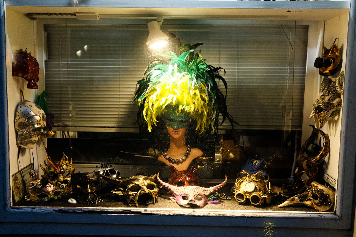 Photo of the display case of a shop with a single spotlight shining on a mannequin bust dressed with a large green, black and yellow feathered wig and wearing a sparkly green mask across its eyes. Masquerade masks of various shapes and colors are scattered at the base of the display case and hang from the left and right walls. 