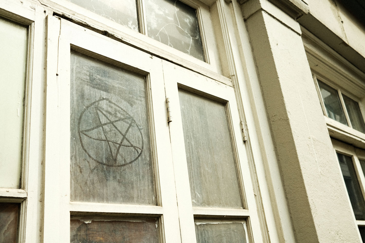 Close up photo of the front of an off-white, dusty beige building with wooden doors. The glass in the doors is very dirty and someone has used their finger to draw a star with a circle around it in the dirty glass.