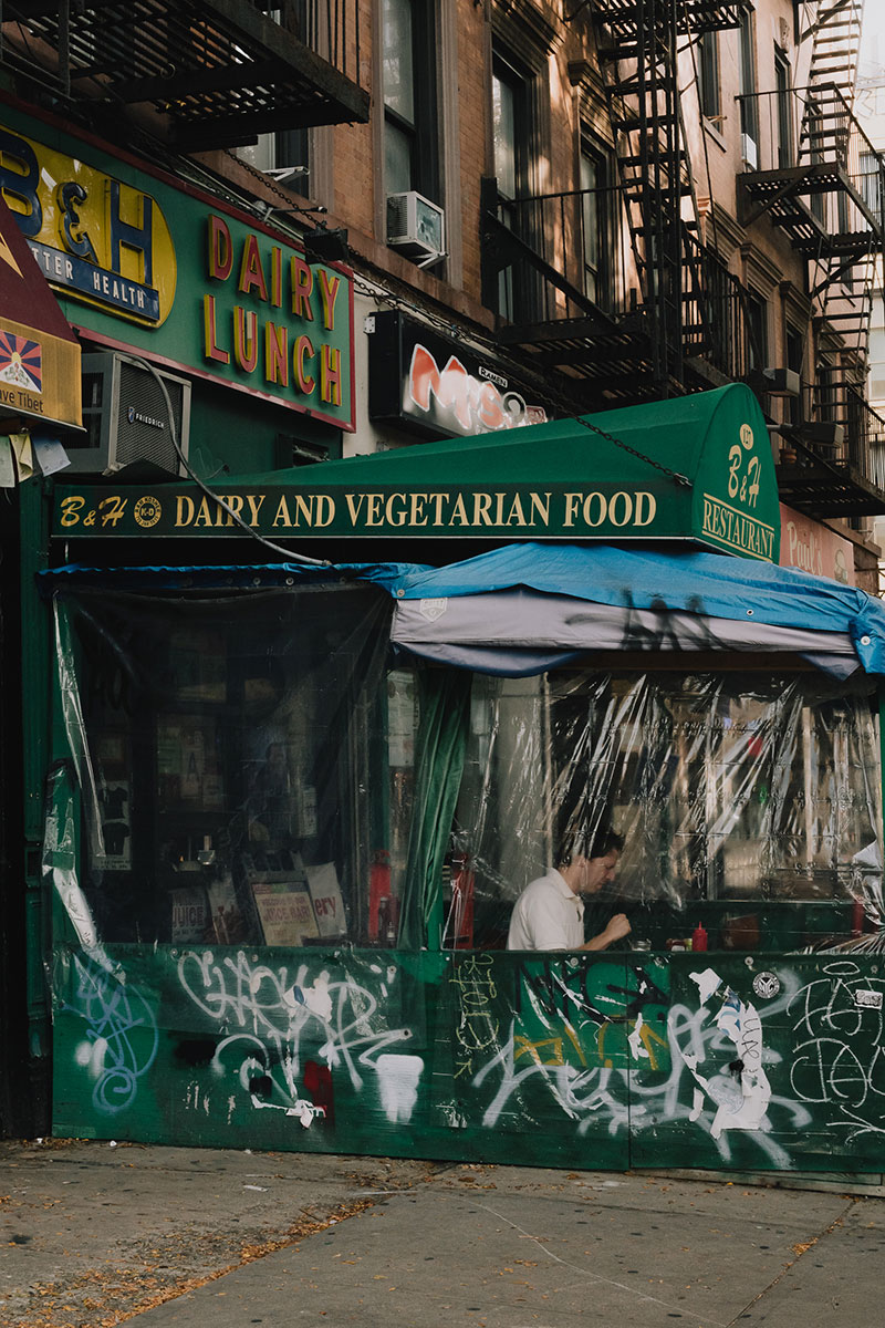 A photo of the side of a diner along a street New York. The diners awning and sign is forest green with red and yellow lettering. An outdoor dining area sealed off with clear plastic insulation is under the awning and a person in a white sheet is eating at a table under it. 