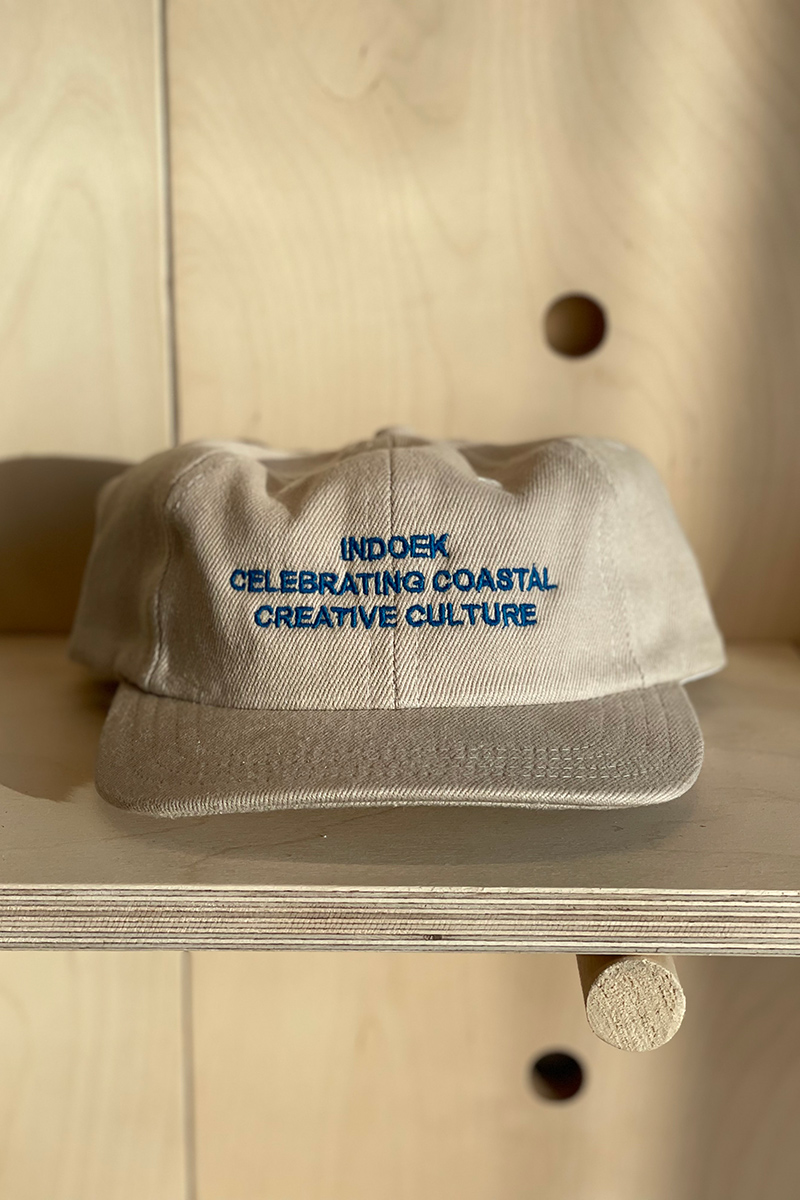 Photo of a khaki hat with navy blue lettering sitting on a light brown wooden display shelf. 