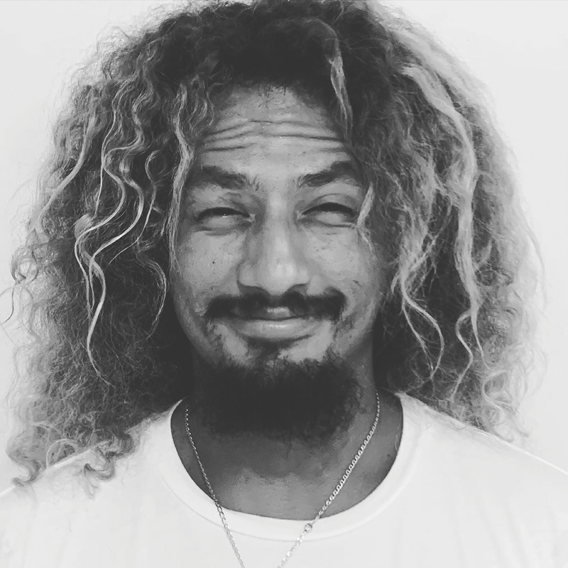 A close-up black and white head shot of a man (Carlos Muñoz) smiling with his lips closed, his eyebrows raised, and his eyes squinting. The man has long, dark, wavy and curly hair with streaks of blonde in it as well as a dark mustache and short beard. He is wearing a white t-shirt and a silver chain around his neck.