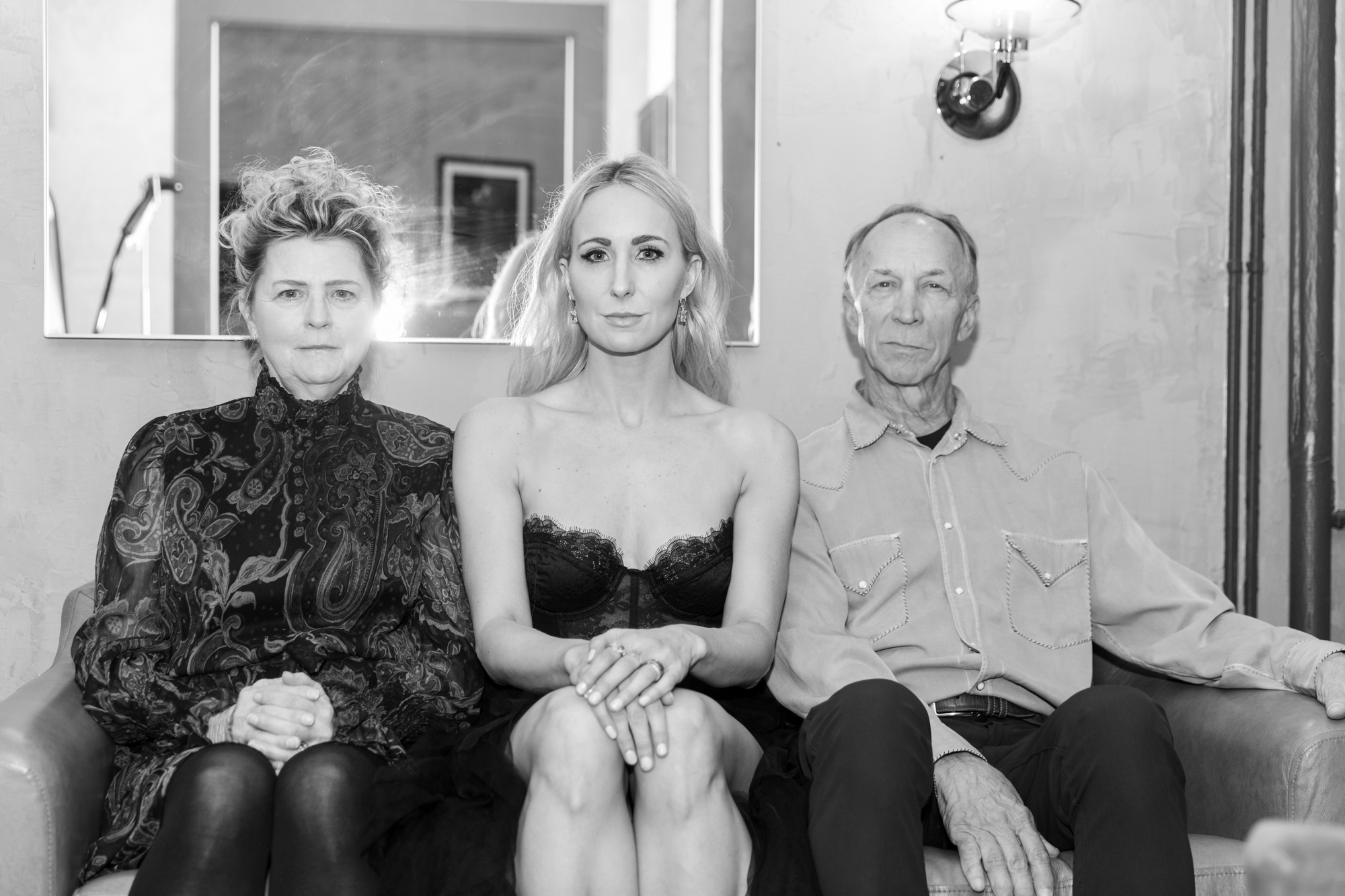 Black and white photo of two women (Nikki Glaser in center) in fancy clothing and a man in a button up shirt all sitting on a couch next to each other in a row and staring at the camera with slight smirks on their faces. There is a mirror hanging on the wall behind them. 