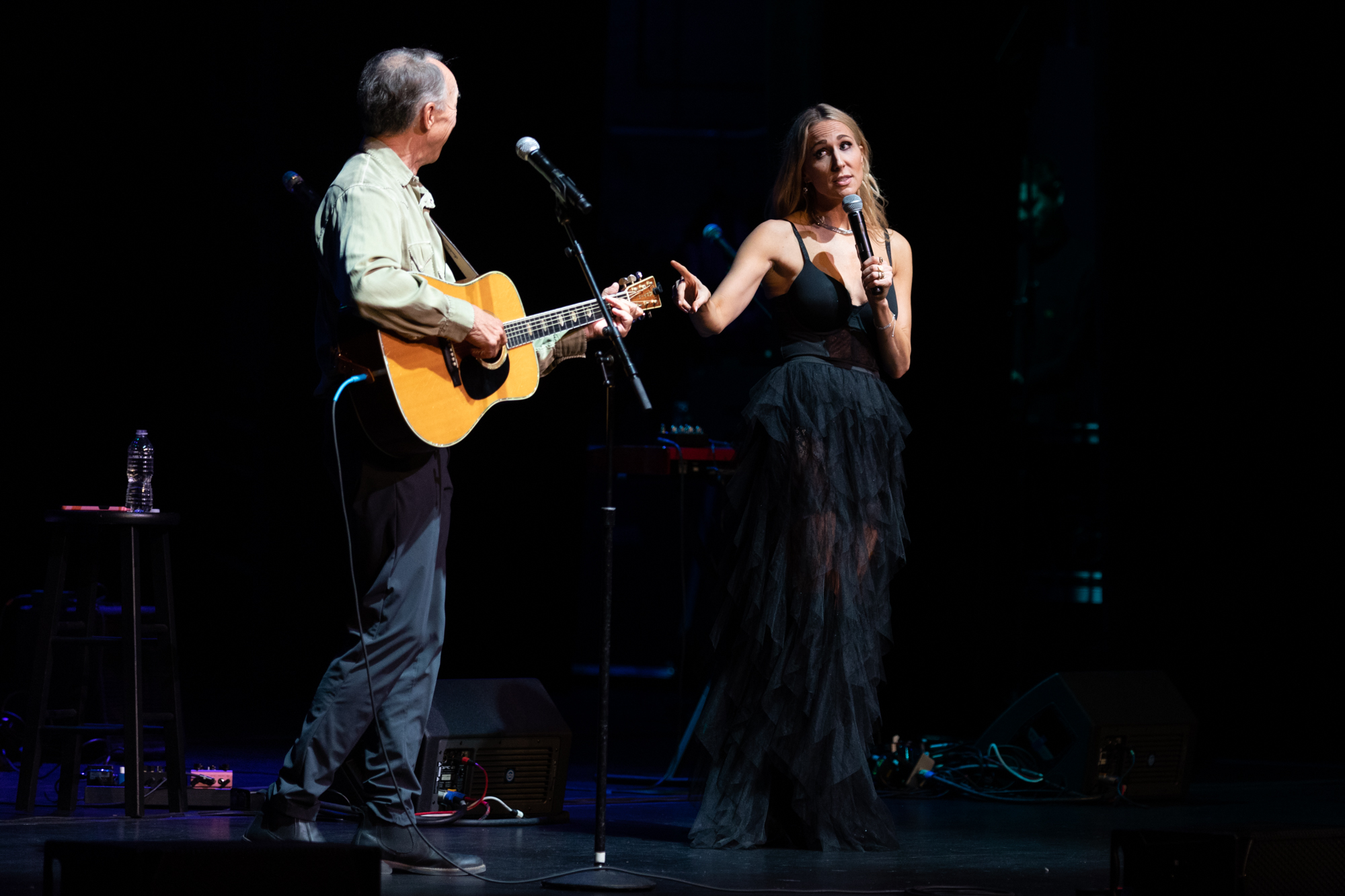 A woman (Nikki Glaser) and a man (Nikki Glaser's dad) standing on a dimly lit black stage. The woman is wearing a long, sheer black dress and speaking into a microphone while pointing at the man to her left. The man is playing a light brown acoustic guitar and looking at the woman. 