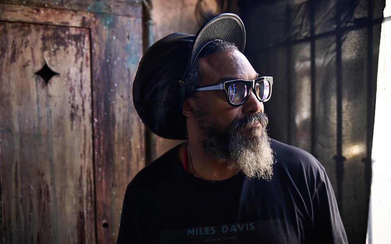 Horizontal portrait of the top half of a gray-bearded man wearing a black t-shirt, a black rastacap and a pair of chunky black glasses with white arms. The man is staring out a window to the the right of the camera. The walls behind him are made of brown battered wood. 