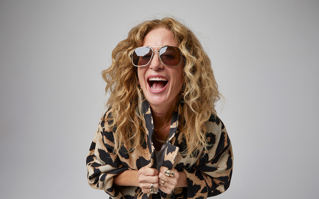 A horizontal portrait of a blonde, wavy haired woman wearing a pair of large gold rimmed and brown tinted aviators. The woman is in the center of the image smiling with her mouth open and clutching her leopard print collared jacket around herself. The background of the image is slate gray. 