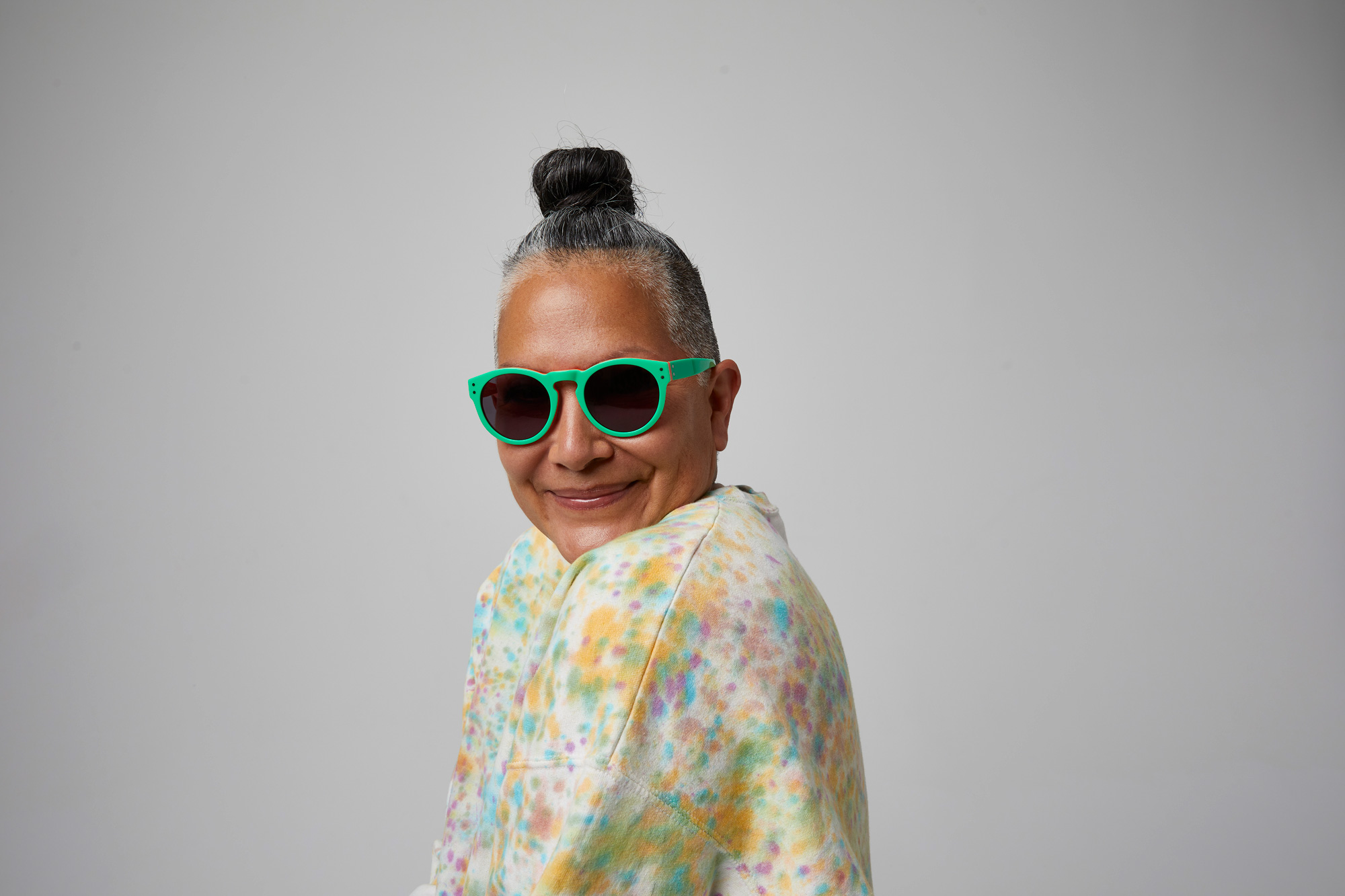 Horizontal portrait of a gray-haired woman wearing her hair in a high bun on the top of her head. The woman's body is facing the side and she is turning her head to look at the camera with a wide smile. She is wearing a multi-colored floral pastel blouse and a pair of bright jade green sunglasses with dark round, gray tinted lenses. The background of the image is light gray.
