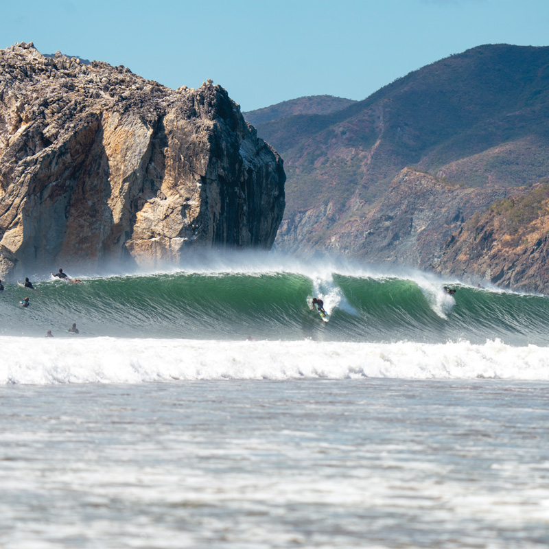 Photo taken from the shore of a teal wave breaking along a rugged, cliff-lined coastline. A man (Carlos Muñoz) is surfing in the center of the wave from the crest to the trough and skimming the water with his hand. Another man is further down to the right of Carlos Muñoz and is just taking off to surf in the other direction. A group of surfers are paddling on their boards past the wave to the left of the photo.