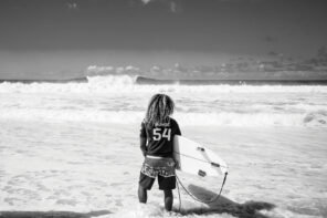 Black and white hero image of the back of a man (Carlos Muñoz) with long curly hair standing in the white water on the shore of a beach holding a surfboard while staring out at the large breaking waves in the distance. The man is wearing a pair of striped board shorts and a dark colored jersey-style rashguard with "54" and "Muñoz" printed in white.