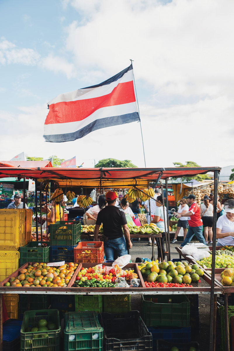 Costa Rican flag flying over the market or bolsa full of stands, fruit, produce, and people.