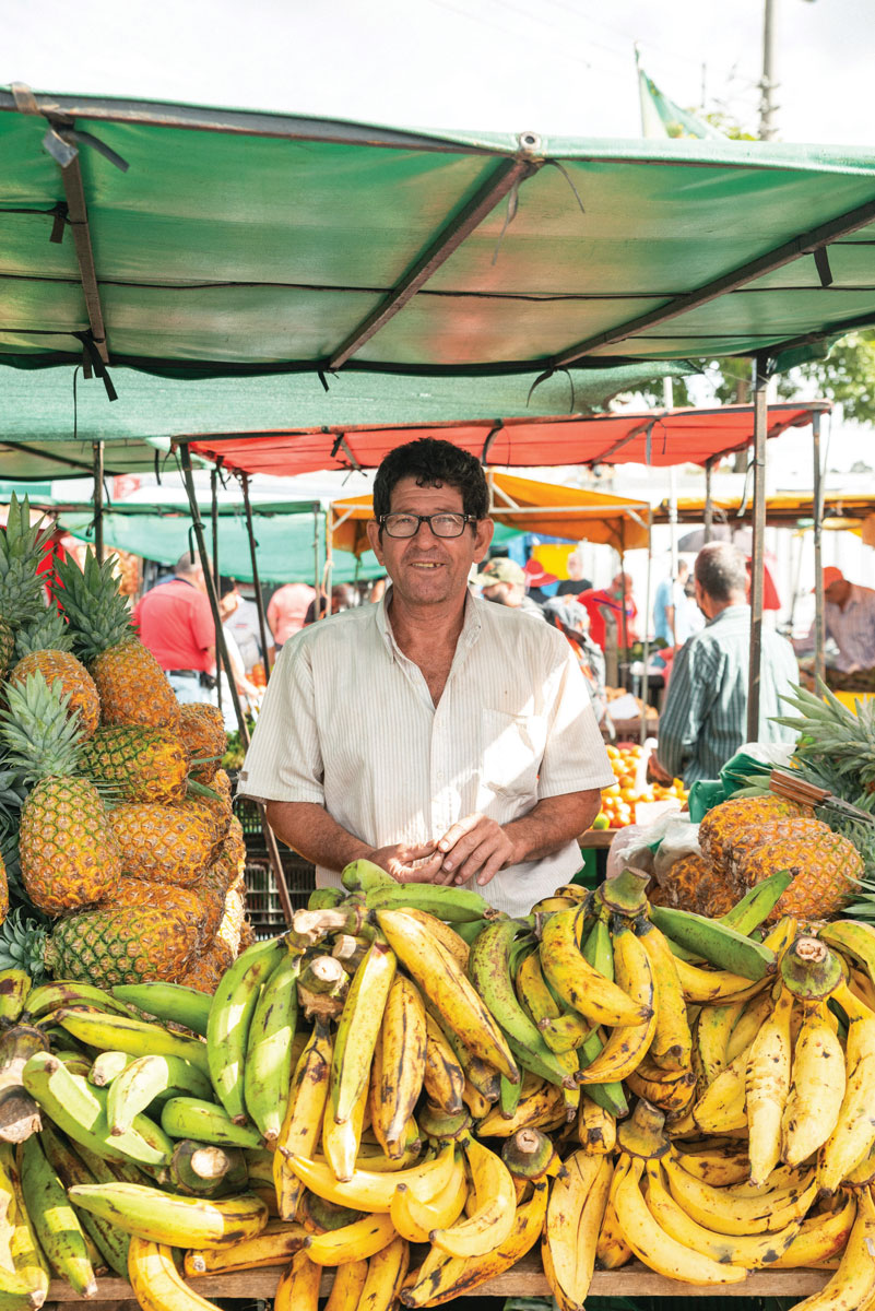 Man smiling and standing behind a stacks plantains and piles of pineapples.