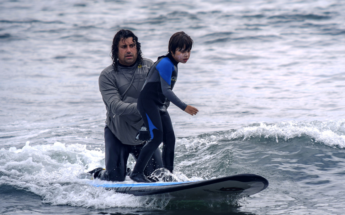 A man with long, curly brown hair kneeling on a surf board out int he water helping balance a young boy in a black and blue wetsuit standing on the same surf board.