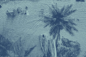 Arial shot of people on a beach with towels. There are upside down wooden boats in the bottom right hand side as well as some scattered palm trees. The image has a blue-colored effect over top of it.
