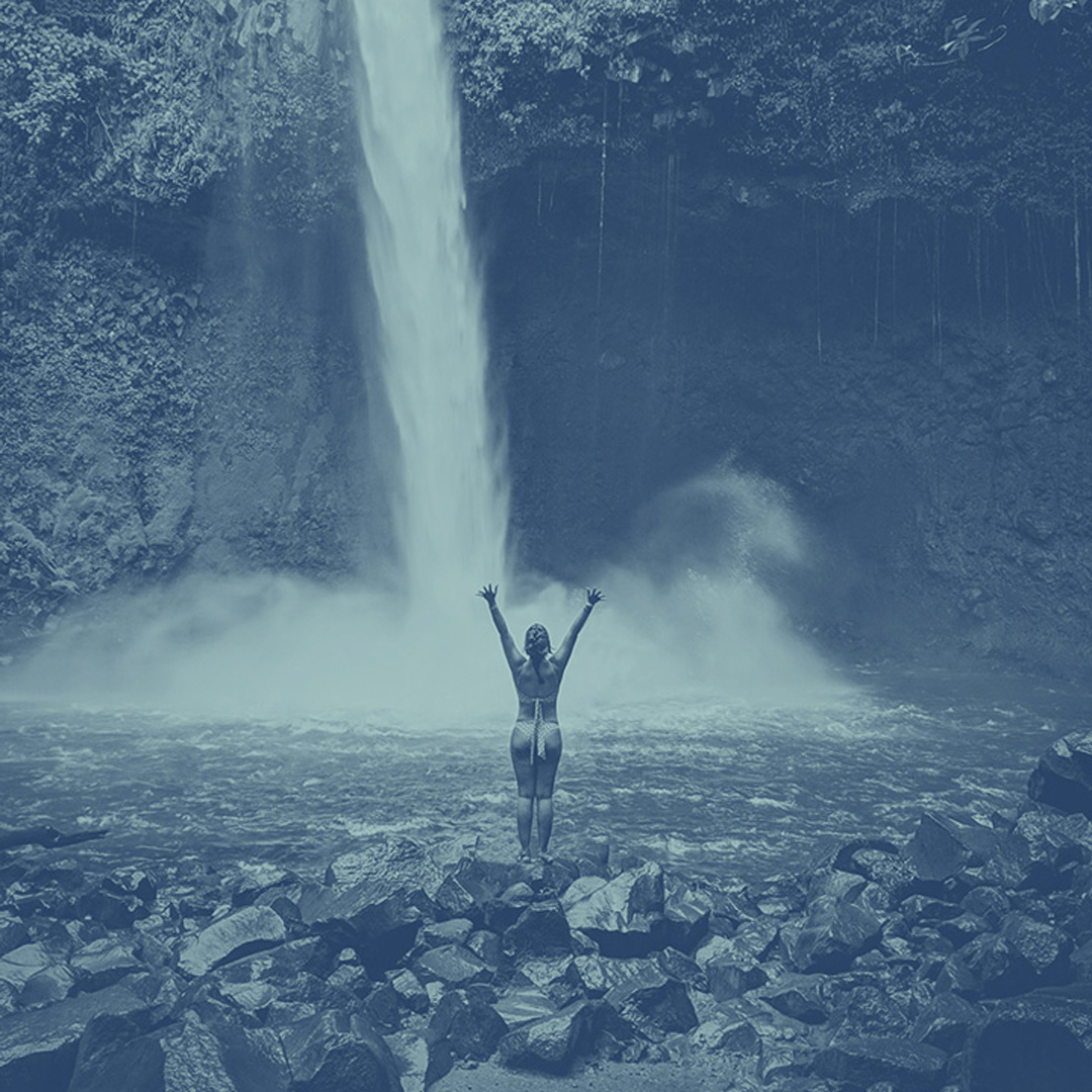 A girl standing with her arms reaching towards the sky standing on rocks in front of a waterfall and pool of water. The Costa Rican jungle is behind the waterfall. The image has a blue-colored effect over top of it.