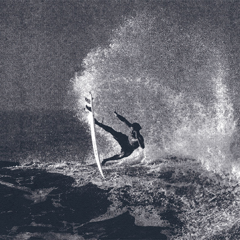 An action shot of a man (Carlos Muñoz) catching some air on his surf board at the crest of a wave in the middle of the ocean. Water is spraying everywhere. The entire image has a dark navy blue overlay on it.