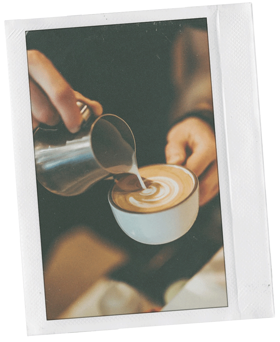 Polaroid photo of someone making latte art. Milk is being poured from a metal cup into a cup of hot coffee in a white mug.
