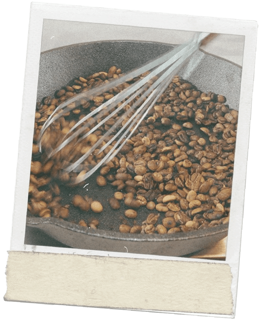 Polaroid photo of a cast iron skillet holding dried coffee beans that are being stirred with a metal whisk. The polaroid has tape along the bottom edge.
