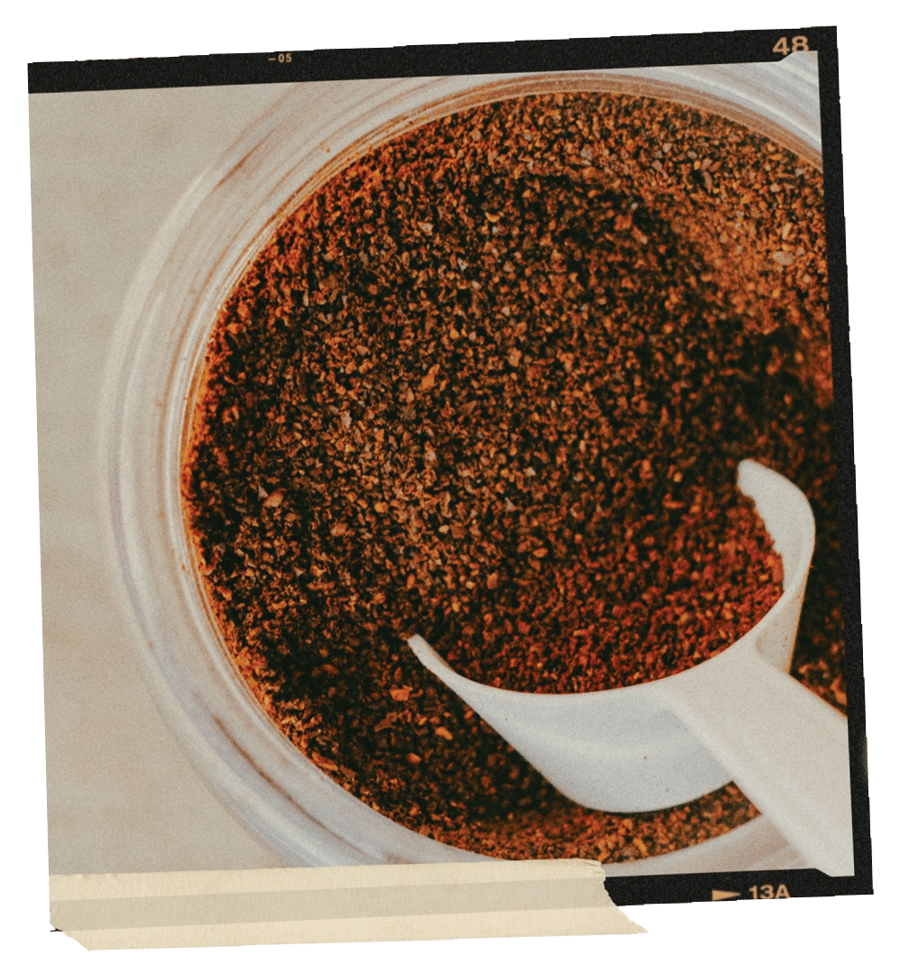 Polaroid photo of a glass jar full of ground coffee with a white scoop in it. The polaroid has tape along the bottom edge.