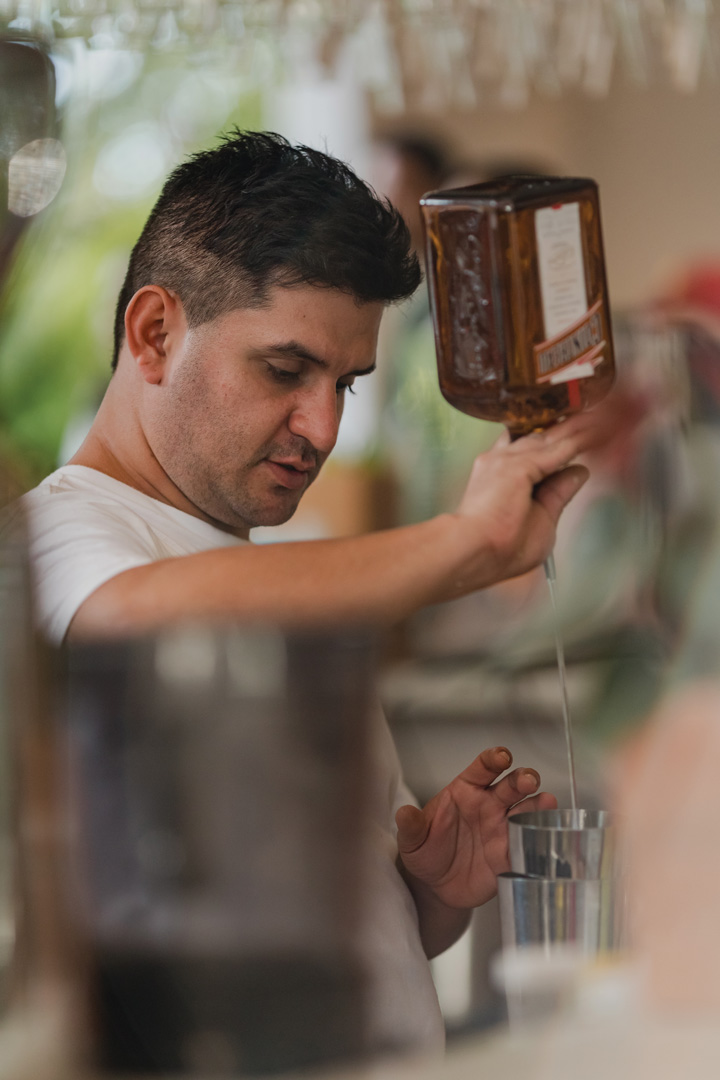 Bartender pouring a brown liquor into a metal shaker. The bartender is wearing a white t-shirt.