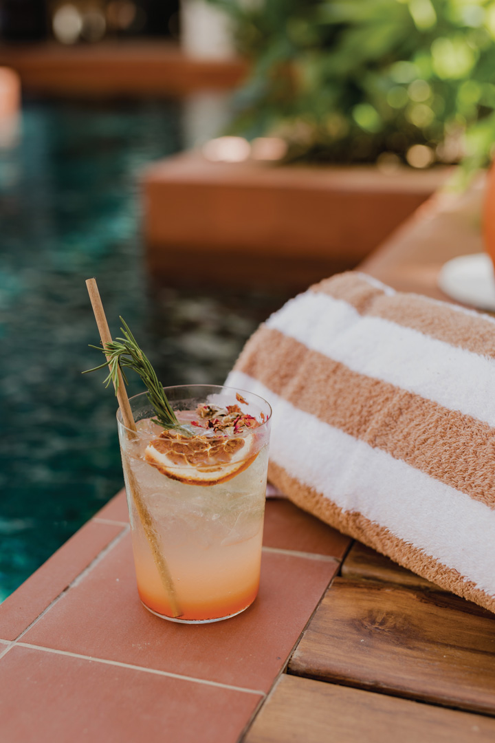 Orange-colored iced cocktail sitting at the edge of the pool. There is an orange straw and dried grapefruit slice in the drink and an orange and white striped towel next to it.