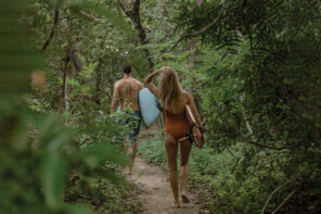 A man and a woman walking away from the camera down a sandy path in the middle of the jungle. They are wearing bathing suits and both carrying surfboards.