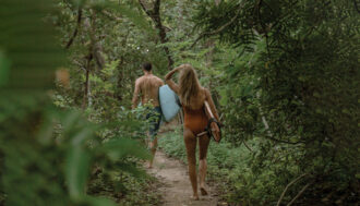 A man and a woman walking away from the camera down a sandy path in the middle of the jungle. They are wearing bathing suits and both carrying surfboards.