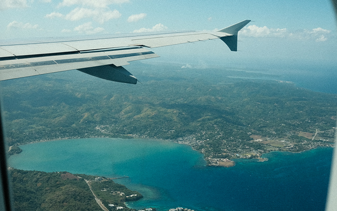 Photo taken from the window seat of an airplane of the the lush, green island of Jamaica surrounded by aquamarine water. The wing of the airplane is visible in the photo to the left.
