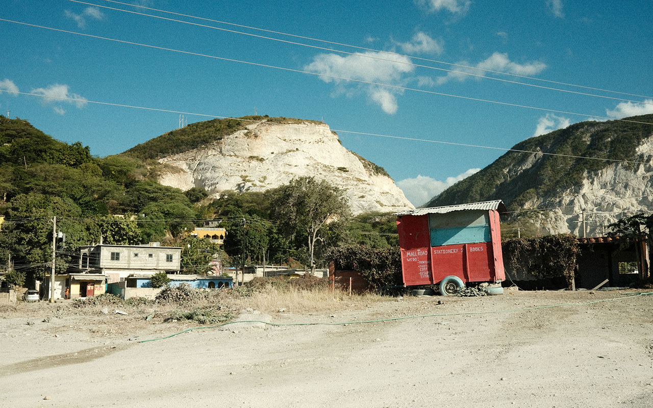 Photo of a dirt road in the chalk white, plant covered mountains of Jamaica. There is an abandoned red wheel cart off the side of the road and a small White House in the background.  