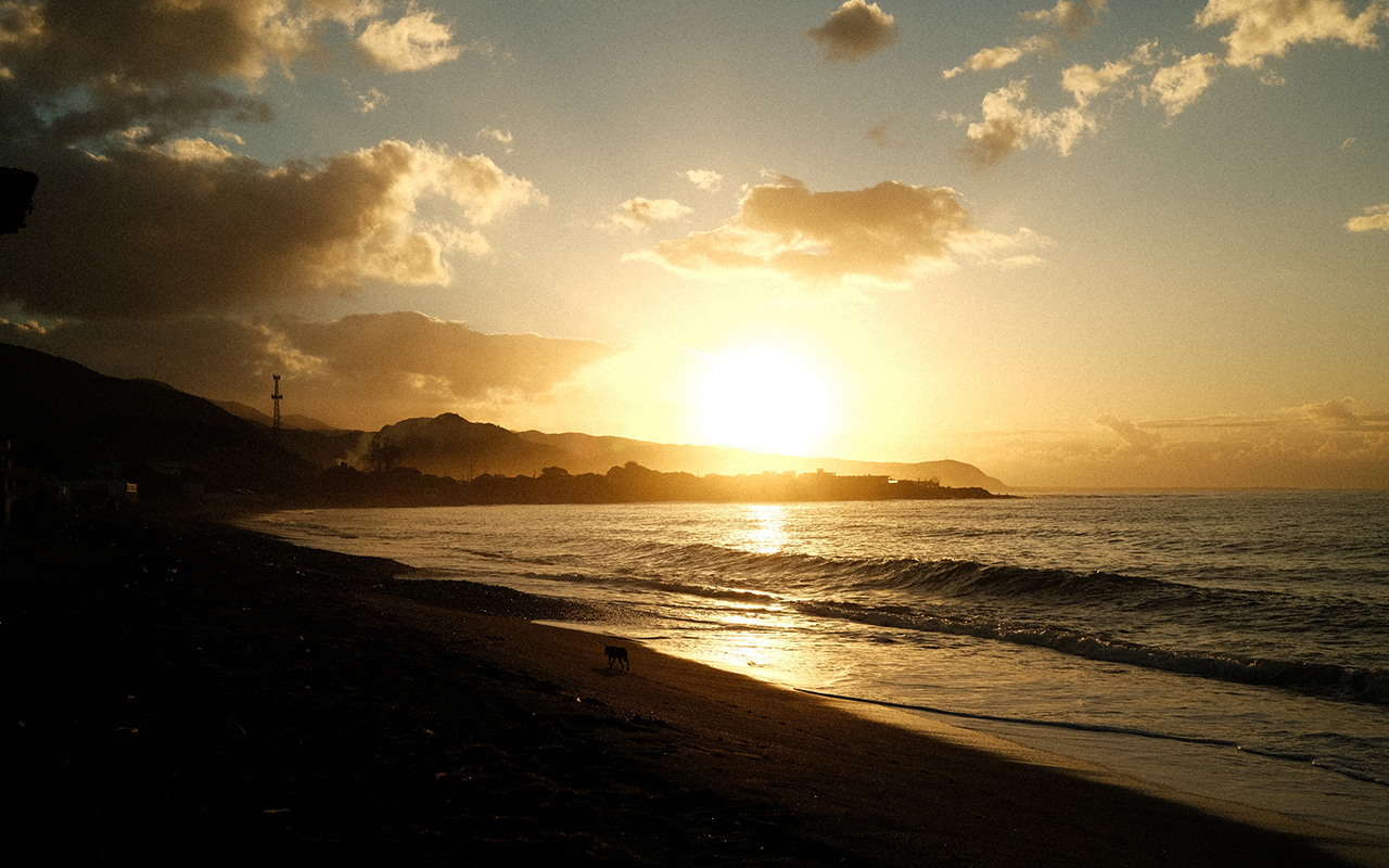 Photo of a light yellow sunset taken from the shores of a beach in Bull Bay. Small, calm waves are lapping the shore and the sun is setting on the mountains in the distance, outlining their dark silhouettes.
