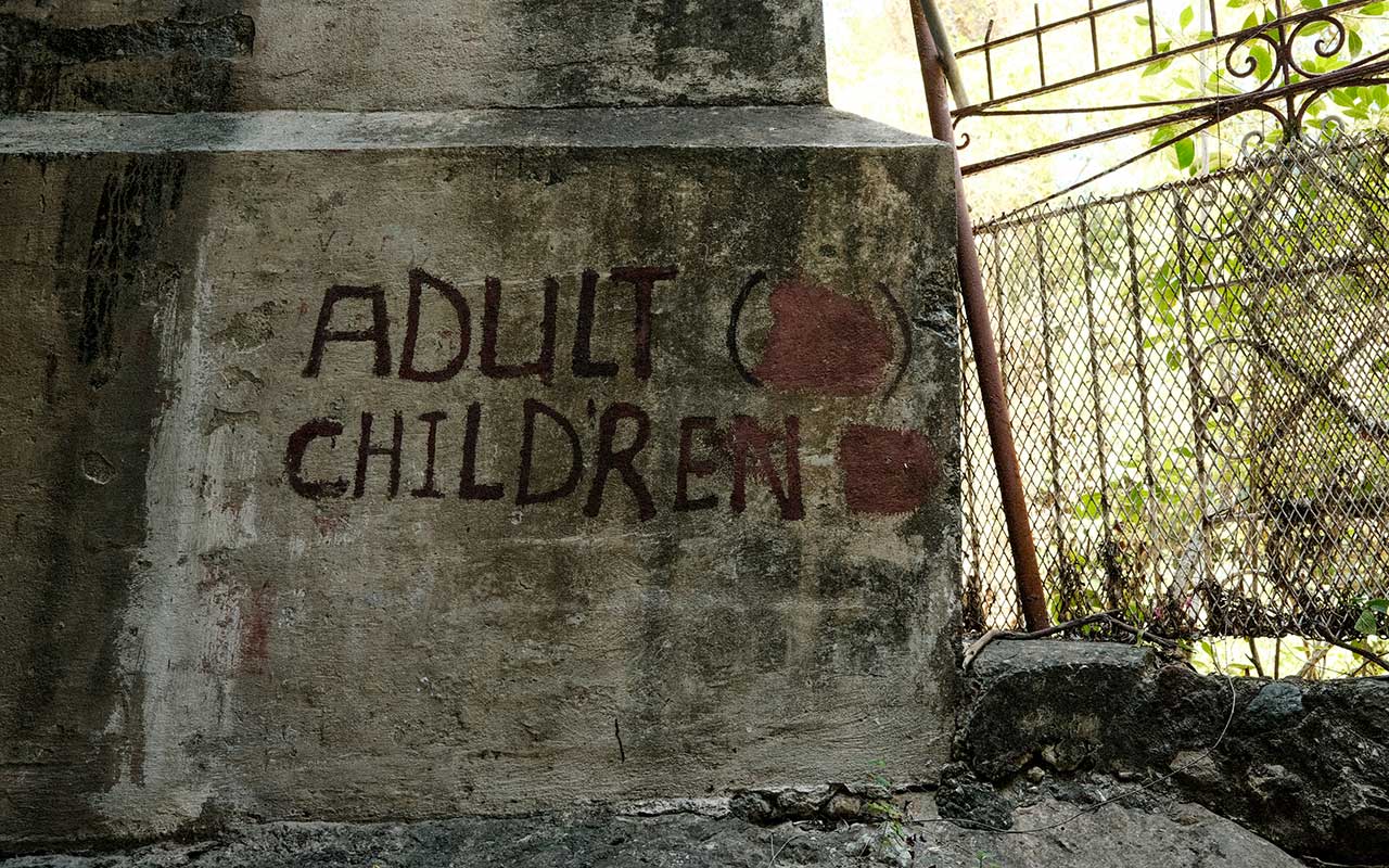 Photo of the base of an old, dirty, dark gray concrete pillar with the word "ADULT CHILDREN" painted in black. An old broken gate and green foliage are visible in the background.