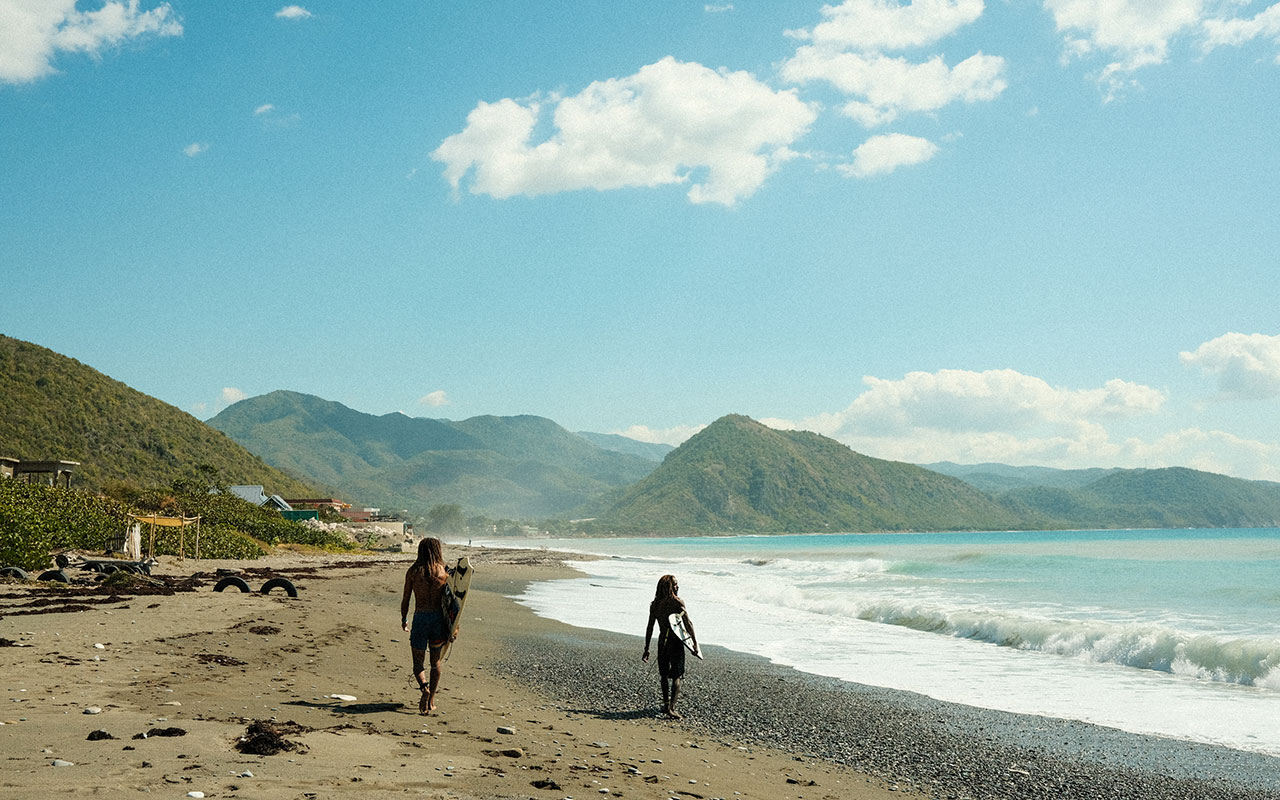 Photo of two long-haired men walking with surfboards under their arms along the sandy and pebbly shore of a beach on a clear day. The sky is light blue with a few clouds. The water is crystal blue and waves are lapping the shore. Lush green mountains are visible in the background.